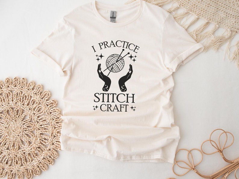 May include: A white t-shirt with black text that reads "I practice stitch craft". The text is surrounded by a graphic of two hands holding a ball of yarn and knitting needles.