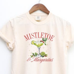 Pu&ograve; includere: Una t-shirt color crema con la grafica di un margarita in un bicchiere con una fetta di lime, bacche di agrifoglio e foglie. Il testo MISTLETOE & MARGARITAS &egrave; stampato in rosso, circondato da piccole stelle dorate.