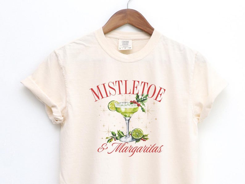 Pu&ograve; includere: Una t-shirt color crema con la grafica di un margarita in un bicchiere con una fetta di lime, bacche di agrifoglio e foglie. Il testo MISTLETOE & MARGARITAS &egrave; stampato in rosso, circondato da piccole stelle dorate.