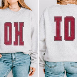 May include: A gray sweatshirt with the word "OHIO" printed in large, burgundy letters on the front and the letters "IO" printed on the back. The sweatshirt is being modeled by a person wearing blue jeans.