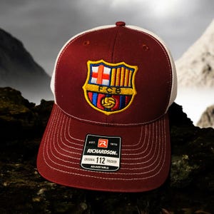 Puede incluir: Una gorra de camionero de color granate y blanco con el logotipo del FC Barcelona bordado en la parte delantera. La gorra es ajustable y tiene una etiqueta de Richardson 112 en el lateral.