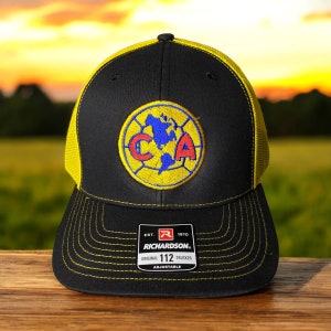 May include: Black and yellow trucker hat with a patch featuring a blue and red emblem. The hat has a curved brim and a mesh back. The patch has the letters "CA" and a map of the Americas. The tag reads "Richardson 112 Trucker".