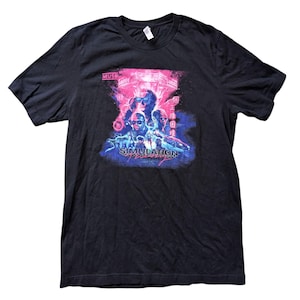 Puede incluir: Camiseta negra con un gráfico vibrante. El diseño incluye el texto "MUSE" en la parte superior y "SIMULATION THEORY" en la parte inferior. El gráfico presenta figuras en tonos azules, rosas y morados, con un diseño futurista.