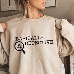 May include: A sand-colored sweatshirt with the text "BASICALLY A DETECTIVE" and a magnifying glass with the letter "A" inside.