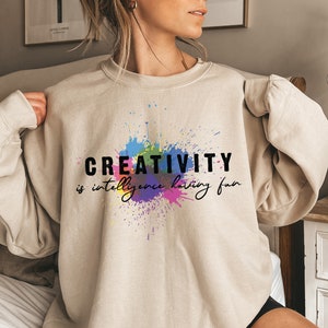 May include: Sand colored sweatshirt with a colorful paint splatter design and the text "CREATIVITY is intelligence having fun".