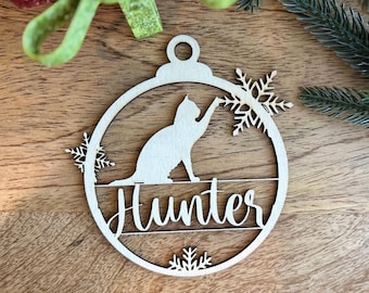May include: A circular light-colored wooden Christmas ornament featuring a cutout silhouette of a cat reaching toward a snowflake. The name Hunter is laser-cut in a cursive script across the center, with additional snowflake accents at the edges.