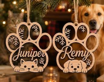 May include: Two light-colored wooden paw-shaped ornaments hanging from twine. The left ornament features the name Juniper above a cat face. The right ornament features the name Remy above a dog face. Both include snowflake and branch cutouts.