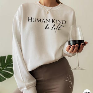 May include: White crew neck sweatshirt with the text "HUMAN KIND be both" in black. The person is wearing brown leggings and holding a wine glass with red wine. A gold necklace is visible.