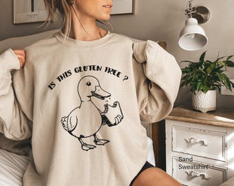 May include: Sand-colored sweatshirt with the text "IS THIS GLUTEN FREE ?" above a cartoon duck eating bread. The duck is black and white. The sweatshirt is a neutral color.