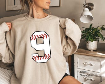 May include: A sand-colored crewneck sweatshirt featuring a large graphic of the number nine. The number is designed with a white fill, a thick black outline, and red stitching details that mimic the appearance of a baseball seam.