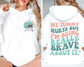May include: A white hooded sweatshirt featuring a teal cartoon character and the text Tummy Ache Survivor Emily on the front. The back displays the text MY TUMMY HURTS BUT I'M BEING REALLY BRAVE ABOUT IT in large, wavy, pink and teal lettering.