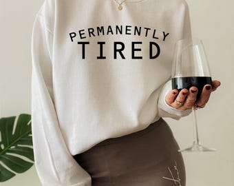 May include: A white long-sleeve crewneck sweatshirt featuring the black text PERMANENTLY TIRED printed across the chest. The garment has a standard fit with ribbed cuffs and a ribbed neckline, providing a clean and simple look for casual wear.