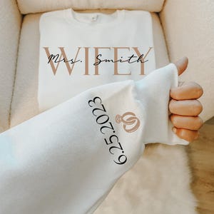 May include: A white long-sleeve sweatshirt featuring the text WIFEY in large tan letters with Mrs. Smith written in black script over it. The sleeve displays the date 6.25.2023 in black text next to a tan graphic of two interlocking wedding rings.