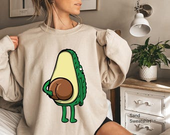 Op de afbeelding: Een zandkleurige sweater met ronde hals en een grafische illustratie van een groene avocado met een bruine pit die zo is geplaatst dat deze op billen lijkt. De avocado heeft dunne armen en benen, een lichtgeel midden en een getextureerde groene schil.
