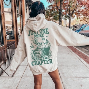 May include: A cream-colored hooded sweatshirt featuring a green graphic on the back. The design displays a skeleton reading a book, surrounded by flowers and butterflies, with the text "JUST ONE MORE CHAPTER" printed in a bold, green font.