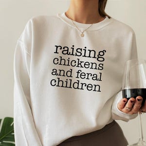 White long-sleeve crewneck sweatshirt featuring the black text raising chickens and feral children printed in a simple serif font across the chest