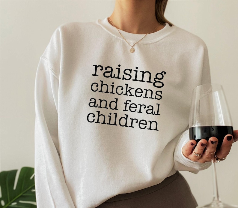 White long-sleeve crewneck sweatshirt featuring the black text raising chickens and feral children printed in a simple serif font across the chest
