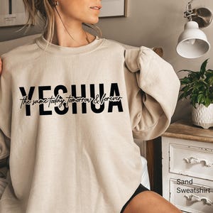 Puede incluir: Sudadera color arena con la palabra "YESHUA" en letras negras en negrita y la frase "the same today, tomorrow & forever" en una fuente cursiva. La sudadera es de color neutro y tiene mangas largas.
