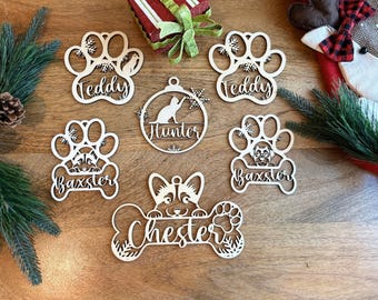 May include: A collection of six laser-cut wooden ornaments featuring pet-themed designs. Four ornaments are shaped like paw prints with names Teddy and Baxster, one is a circular bauble with a cat and the name Hunter, and one is a bone shape with the name Chester.