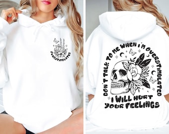 Puede incluir: Una sudadera con capucha blanca con un gráfico negro en el pecho de una mano de esqueleto haciendo el signo de la paz con flores y la palabra OVERSTIMULATED. La espalda muestra una calavera con flores, mariposas y el texto DON'T TALK TO ME WHEN I'M OVERSTIMULATED I WILL HURT YOUR FEELINGS.
