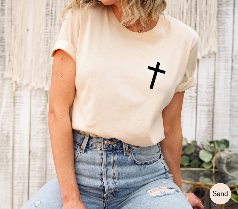 Sand-colored short-sleeve t-shirt featuring a small, simple black cross graphic printed on the left chest area