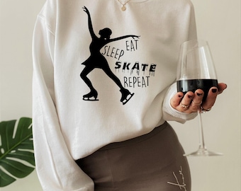 May include: A white long-sleeve sweatshirt featuring a black graphic of a figure skater and the text "SLEEP EAT SKATE REPEAT" printed on the front. The design includes a stylized silhouette of a person skating and bold, dripping-style lettering.