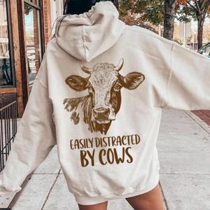 May include: A cream-colored hooded sweatshirt featuring a brown graphic of a cow on the back. Below the cow illustration, the text reads "EASILY DISTRACTED BY COWS" in a bold, brown, sans-serif font. The hoodie has long sleeves and a hood.