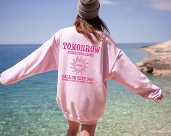 Könnte beinhalten: Ein hellrosa Langarm-Sweatshirt mit dunkelrosa Text auf der Rückseite, der TOMORROW NEEDS YOUR LIGHT, eine Sonnengrafik und CALL OR TEXT 988 NATIONAL SUICIDE PREVENTION LIFELINE lautet. Das Kleidungsstück wird mit einer grauen Mütze getragen.