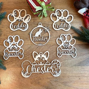 May include: Wooden pet-themed Christmas ornaments with names like "Teddy", "Baxter", "Hunter", and "Chester". The ornaments feature paw prints, dog and cat silhouettes, and a bone shape. Festive holiday decor.