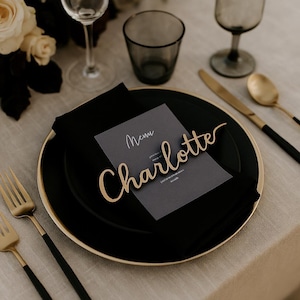 May include: A gold-toned laser-cut name tag featuring the name Charlotte in a cursive script font, placed atop a dark gray menu card, a black linen napkin, and a black dinner plate with a gold rim, accompanied by gold and black cutlery.