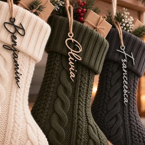 Puede incluir: Tres calcetines navideños de punto trenzado en color crema, verde oliva y gris carbón. Cada calcetín cuenta con una etiqueta de madera colgante con los nombres Benjamin, Olivia y Francesca en letra cursiva, sujetos por cordel.