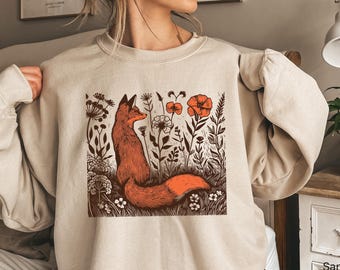 May include: A beige crewneck sweatshirt featuring a square graphic print of a fox sitting among various wildflowers. The fox is rendered in shades of orange and brown, surrounded by detailed botanical illustrations in a dark brown ink style.