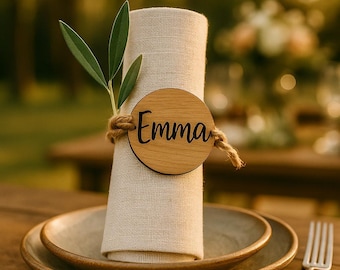 May include: A beige linen napkin rolled into a cylinder, secured with a circular wooden tag featuring the name Emma in black script. A piece of jute twine wraps around the napkin, holding a small green olive branch sprig against the wooden tag.