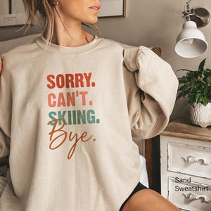 May include: Sand-colored sweatshirt with the text "SORRY. CAN'T. SKIING. Bye." in red, teal, and orange lettering. The long-sleeved crewneck top is a casual style.