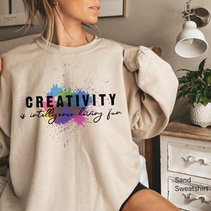 May include: Sand-colored sweatshirt with the words "CREATIVITY is intelligence having fun" in black and script font over a colorful paint splatter design. The sweatshirt is a neutral color.