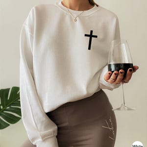 White long-sleeve crewneck sweatshirt featuring a small black cross graphic on the upper left chest