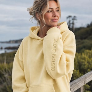 Puede incluir: Una sudadera con capucha de color amarillo pálido con mangas largas y un bolsillo tipo canguro frontal. El texto CUSTOM QUOTE está bordado verticalmente en hilo amarillo a juego en el lado exterior de la manga izquierda, cerca del puño.