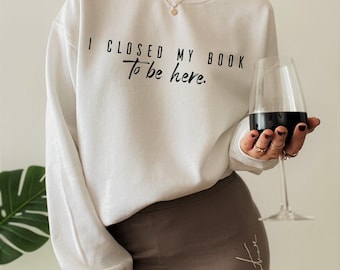 May include: A white long-sleeve crewneck sweatshirt featuring the black printed text I CLOSED MY BOOK To be here. The garment has a standard fit with ribbed cuffs and a ribbed neckline, worn with brown leggings and a gold chain necklace.