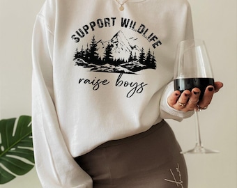 May include: A white long-sleeve sweatshirt featuring a black graphic print of a mountain range and pine trees. The text "SUPPORT WILDLIFE" is printed in a bold, distressed font above the graphic, with the words "raise boys" in script below.