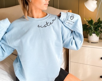 May include: A light blue crewneck sweatshirt featuring the word "sister" in black cursive script on the upper chest. The left sleeve displays a black graphic of a heart topped with a crown, accompanied by the text "emmy alex" in cursive script.