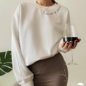 May include: White crew neck sweatshirt with the script "I will Not be Shaken" on the upper left chest. The person is wearing brown leggings and holding a wine glass with red wine.