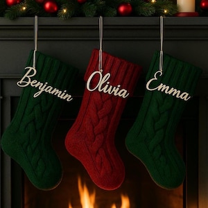 May include: Three cable-knit Christmas stockings hang from a dark mantel. Two are green, and one is red. Each stocking has a name tag: "Benjamin," "Olivia," and "Emma." The background includes a fireplace and Christmas decorations.