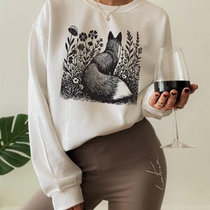 May include: A white long-sleeve sweatshirt featuring a black graphic print of a fox sitting in a field of wildflowers. The sweatshirt has a crew neckline and long sleeves. The graphic is centered on the chest and depicts a fox facing away.