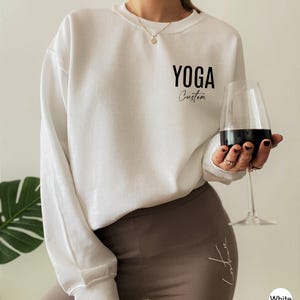 May include: A white long-sleeve crewneck sweatshirt featuring the black text YOGA and the word Custom in a cursive script on the upper left chest. The sweatshirt is paired with solid brown leggings that have a small white script logo on the hip.