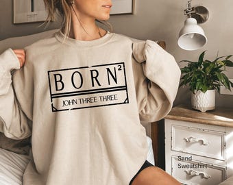 May include: A sand-colored crewneck sweatshirt featuring a black rectangular graphic on the chest. The design includes the text BORN squared and the words JOHN THREE THREE inside the box, with a distressed, weathered texture on the border.