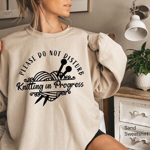 May include: A sand-colored crewneck sweatshirt featuring black text that reads "PLEASE DO NOT DISTURB" arched over a graphic of a ball of yarn with two knitting needles. Below the yarn, the text reads "Knitting in Progress" in script font.