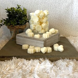 Puede incluir: Una colecci&oacute;n de ceras arom&aacute;ticas de color marfil en varias formas, incluyendo flores y corazones. Algunas ceras est&aacute;n en un tarro de cristal transparente, mientras que otras est&aacute;n dispersas sobre una superficie de madera. Una peque&ntilde;a planta en maceta est&aacute; en el fondo.