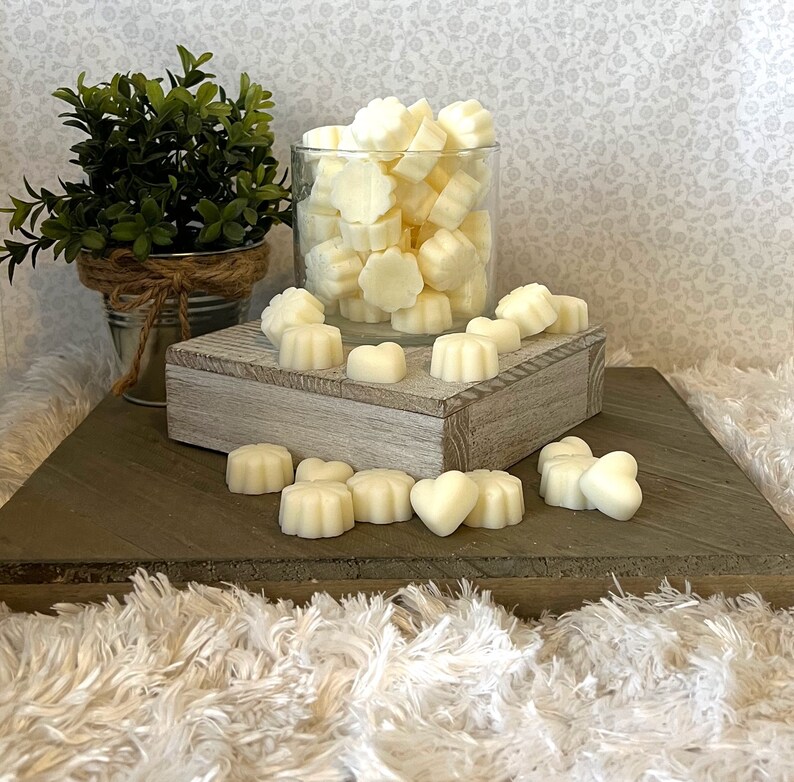 Puede incluir: Una colecci&oacute;n de ceras arom&aacute;ticas de color marfil en varias formas, incluyendo flores y corazones. Algunas ceras est&aacute;n en un tarro de cristal transparente, mientras que otras est&aacute;n dispersas sobre una superficie de madera. Una peque&ntilde;a planta en maceta est&aacute; en el fondo.