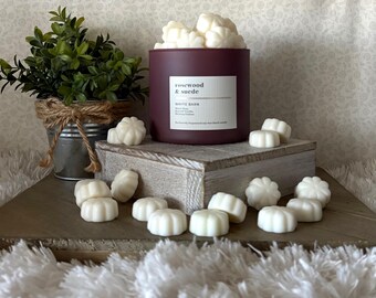 May include: A burgundy candle with the text "rosewood & suede" and "White Barn" on a white label. The candle is filled with white wax melts. A small potted plant and additional wax melts are also present.