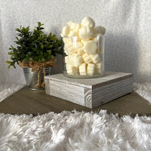 Puede incluir: Un tarro de cristal transparente lleno de ceras perfumadas de color marfil con forma de flor. El tarro est&aacute; sobre un bloque de madera blanqueada. A la izquierda hay una peque&ntilde;a planta en maceta en un cubo de metal galvanizado. El fondo es una tela con estampado floral.